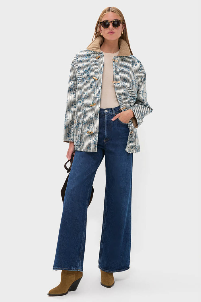 A person wearing Hunter Bell sunglasses, an oversized barn jacket, beige top, blue Italian stretch cotton denim wide-leg jeans, and brown heeled boots stands holding a dark-colored handbag.