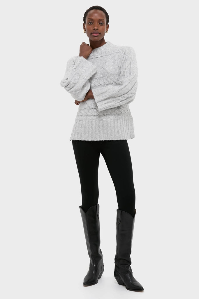 Woman in a classic Gray Cableknit Gladwyn Sweater with a mock neck and wide sleeves, paired with black leggings and knee-high boots for an effortless, high-end look.
